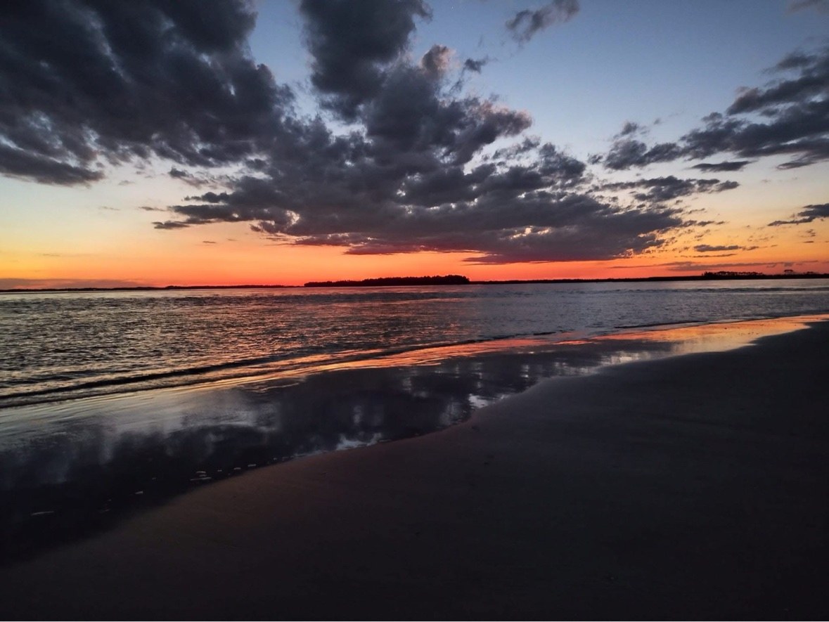 A burning orange dawn sky and dark sculpted clouds mirrored across wet sand